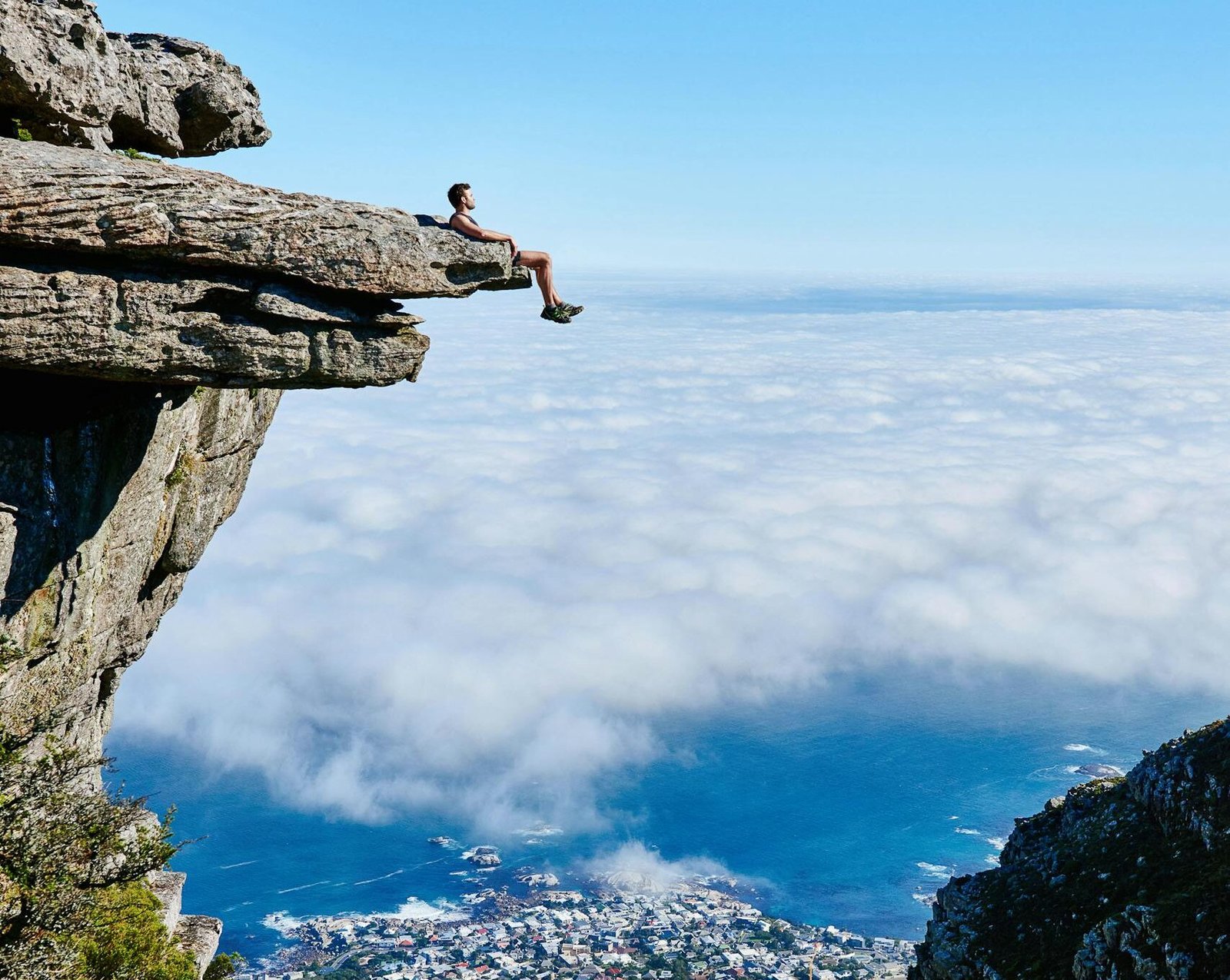 A person sits on a cliff edge enjoying a breathtaking view of the ocean and city below.