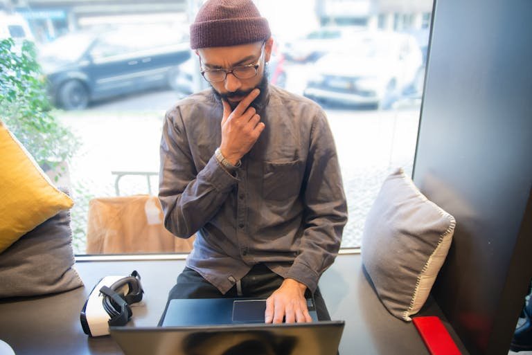 Focused man using laptop in a cozy coffee shop with a VR headset beside him.